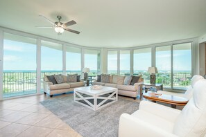 A spacious living room with a large window, a ceiling fan, and a view of the ocean. - Three Bedroom with Media Room with Wrap-Around Balcony. Complimentary Boat Slip! (Orange Beach)