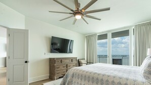 Un dormitorio con cama, ventilador de techo, televisor, cómoda y vista al cielo a través de amplias ventanas.