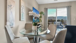 Una sala de estar moderna con una mesa y sillas de vidrio, un cuadro grande, una planta en maceta y vistas al exterior.