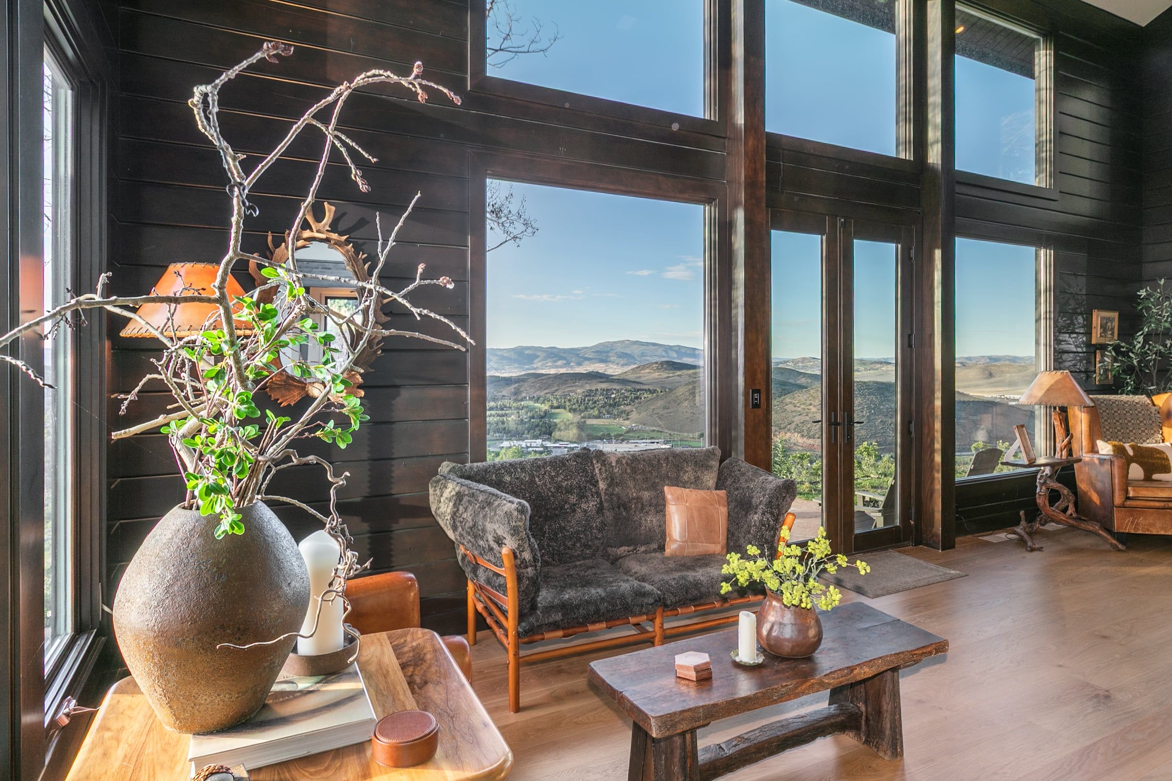 A room with a large window offering a view of a mountainous landscape, a stone vase with branches, a dark sofa, and a wooden coffee table.