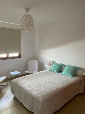 A bedroom with a large bed, a chair, a small table, and a window with blinds.