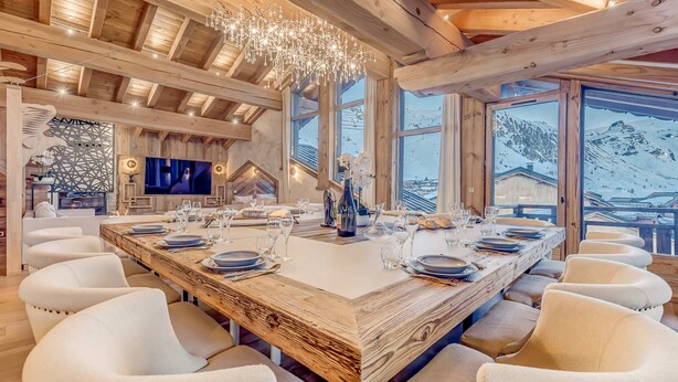 A spacious dining area with a long wooden table set for a meal, surrounded by white upholstered chairs. - 5* Chalet in Tignes - 450m² - 16 guests - Sauna, Jacuzzi, Game Room (Tignes)