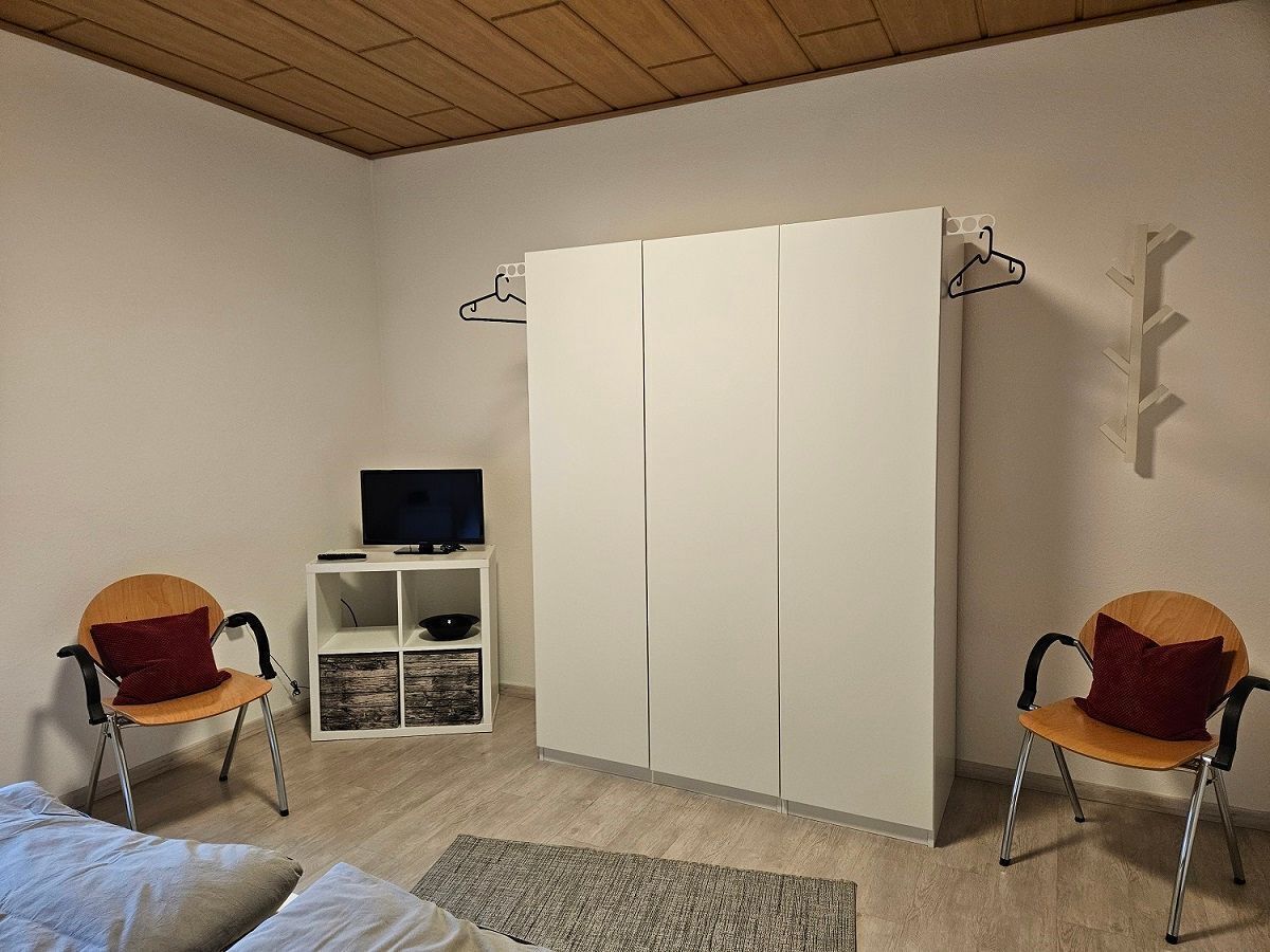 Een slaapkamer met een bed, twee stoelen, een televisie, een kledingkast en een plank met een schaal.