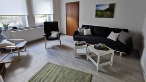 Een moderne woonkamer met een zwarte bank, een houten stoel, een witte salontafel en een groen vloerkleed. Er zijn twee ramen met zonwering, een deur en een schilderij aan de muur.