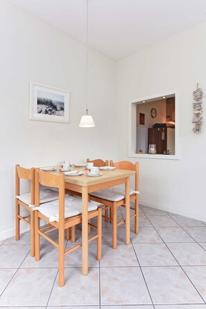 Ein Essbereich mit Holztisch und -stühlen, einer Pendelleuchte, einem gerahmten Bild an der Wand und einem Fenster mit Blick auf einen Küchenbereich.