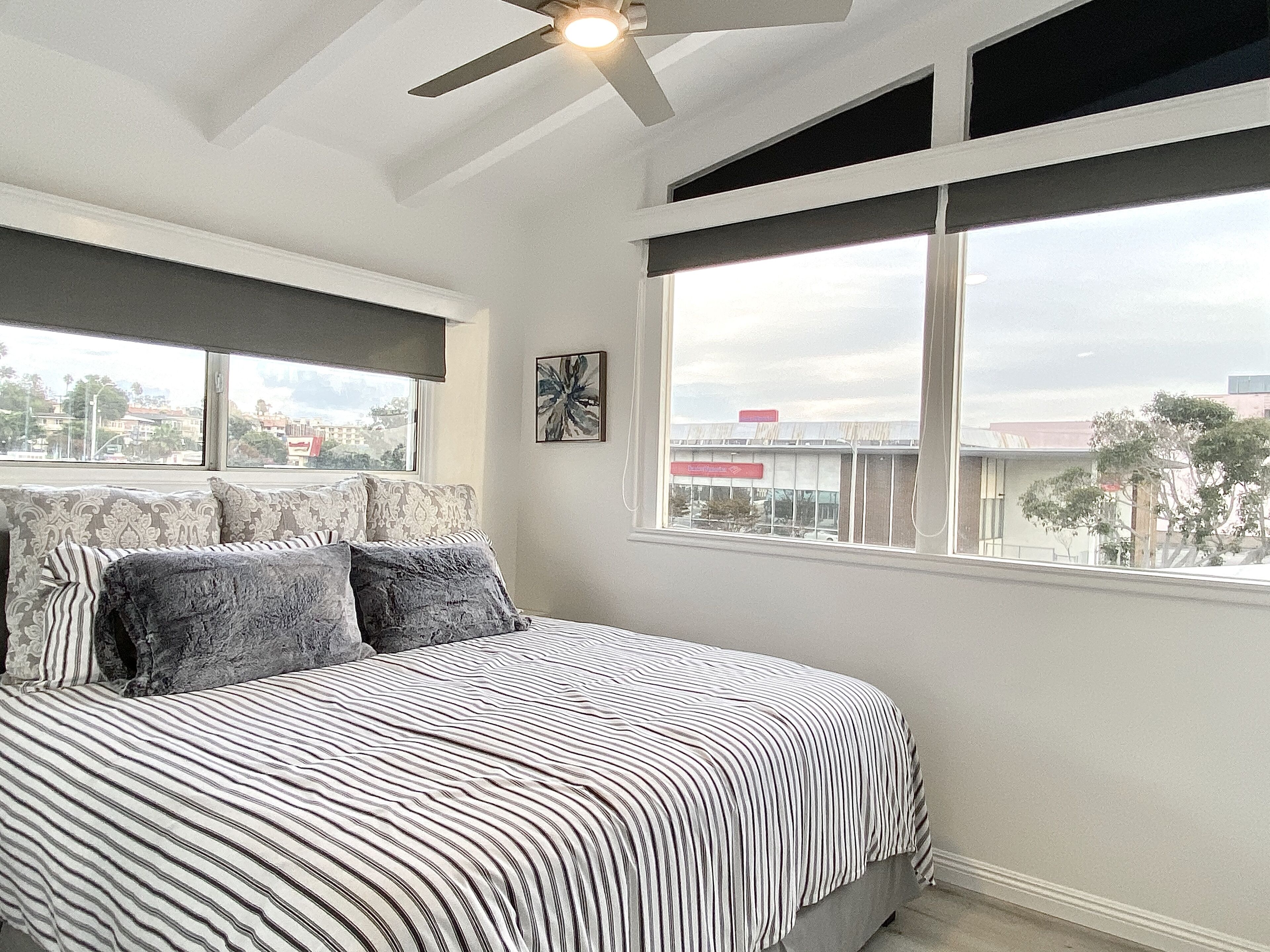 Quarto com cama, ventilador de teto, janela com vista para edifícios e um quadro na parede.