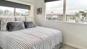 Un dormitorio con cama, ventilador de techo, ventana con vistas a edificios y un cuadro en la pared.