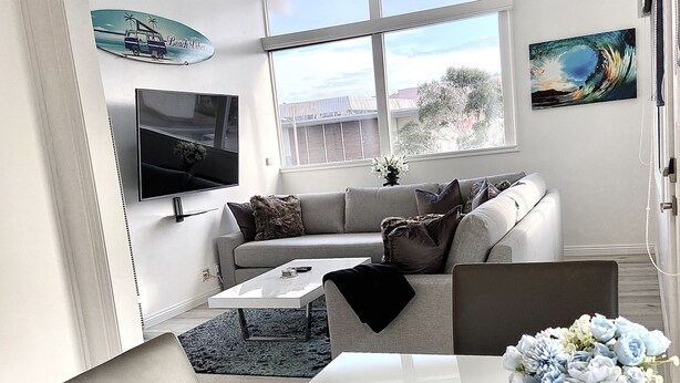 Una sala de estar moderna con un sofá blanco, una mesa de centro y una mesa de comedor. Hay un televisor montado en la pared, un ventilador de techo y un ventanal con vistas al exterior.