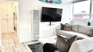 Una sala de estar moderna con un sofá gris, un televisor de pantalla plana montado en la pared y un ventanal con vistas a un edificio.