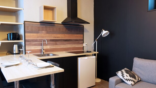 A compact kitchen with a wooden backsplash, a black range hood, a sink, and a compact dishwasher. There is a dining area with a table set for two, and a sofa with a patterned pillow.