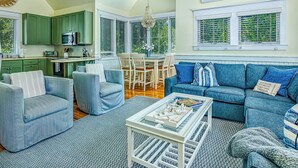 A spacious living room with a blue sofa, a coffee table, and a dining area with a chandelier. - Reverse floor plan cottage with screened in porch. Nature views! (Bald Head Island)