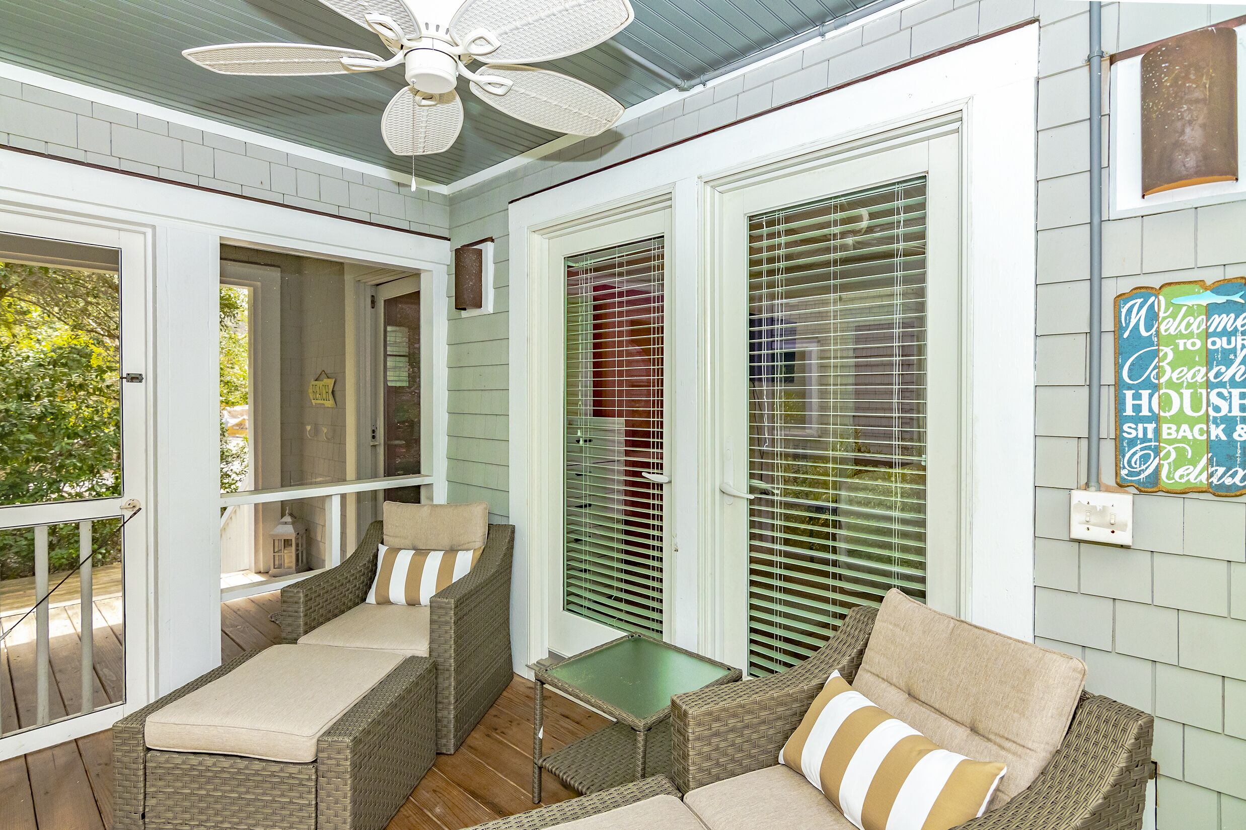 A covered porch with wicker furniture, a ceiling fan, and a sign welcoming guests to the beach house.