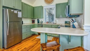 A kitchen with green cabinets, a stainless steel refrigerator, a large island with stools, and a decorative wall piece. - Reverse floor plan cottage with screened in porch. Nature views! (Bald Head Island)