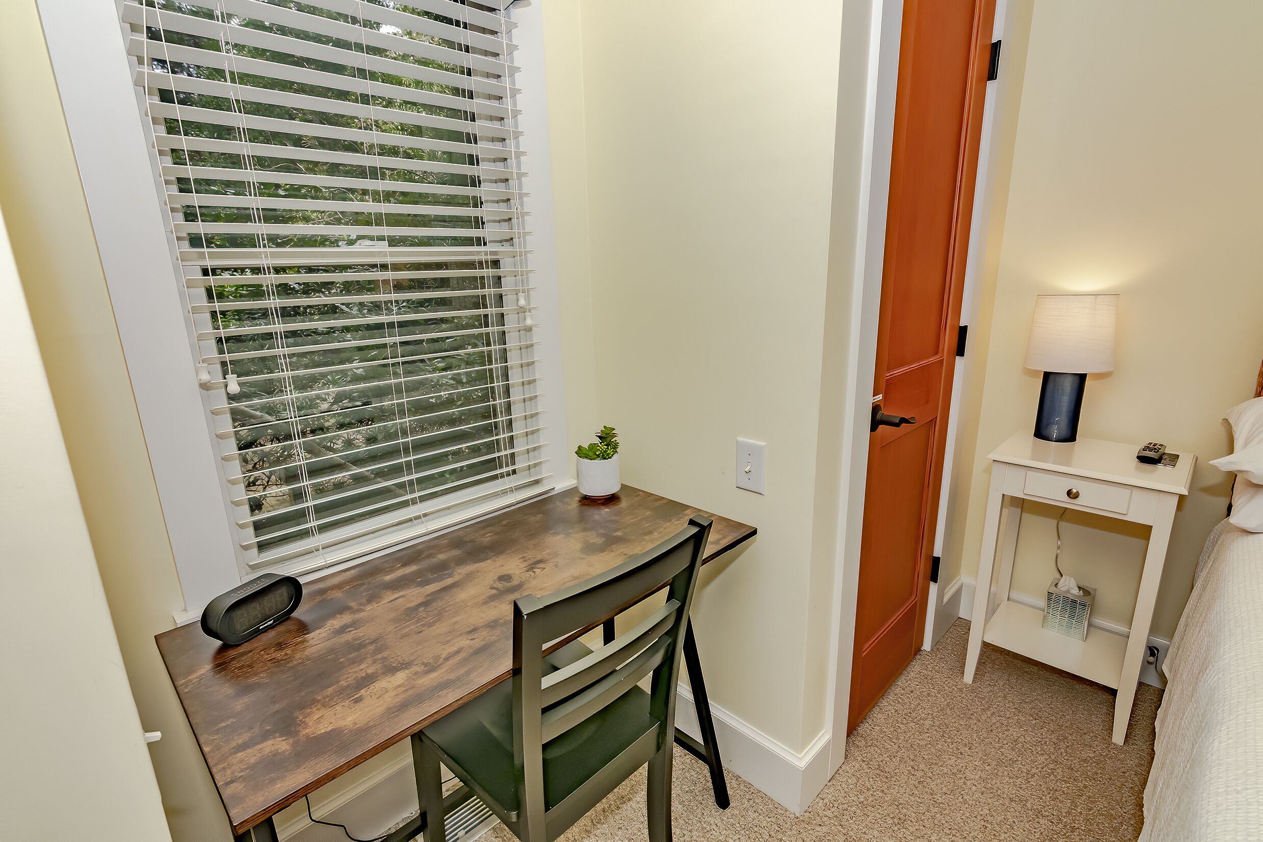 A small, neatly arranged bedroom with a wooden desk, a chair, a bedside table with a lamp, and a window with blinds.