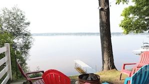 A lakeside view with red and blue Adirondack chairs, a fire pit, and a dock extending into the water.