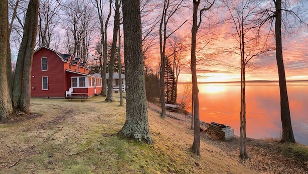 A red cabin with a bench and picnic table by a lake at sunset.