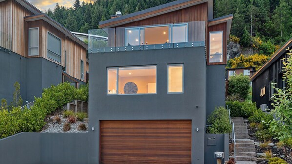 A modern two-story house with a large garage, a balcony, and a view of a forested hillside.