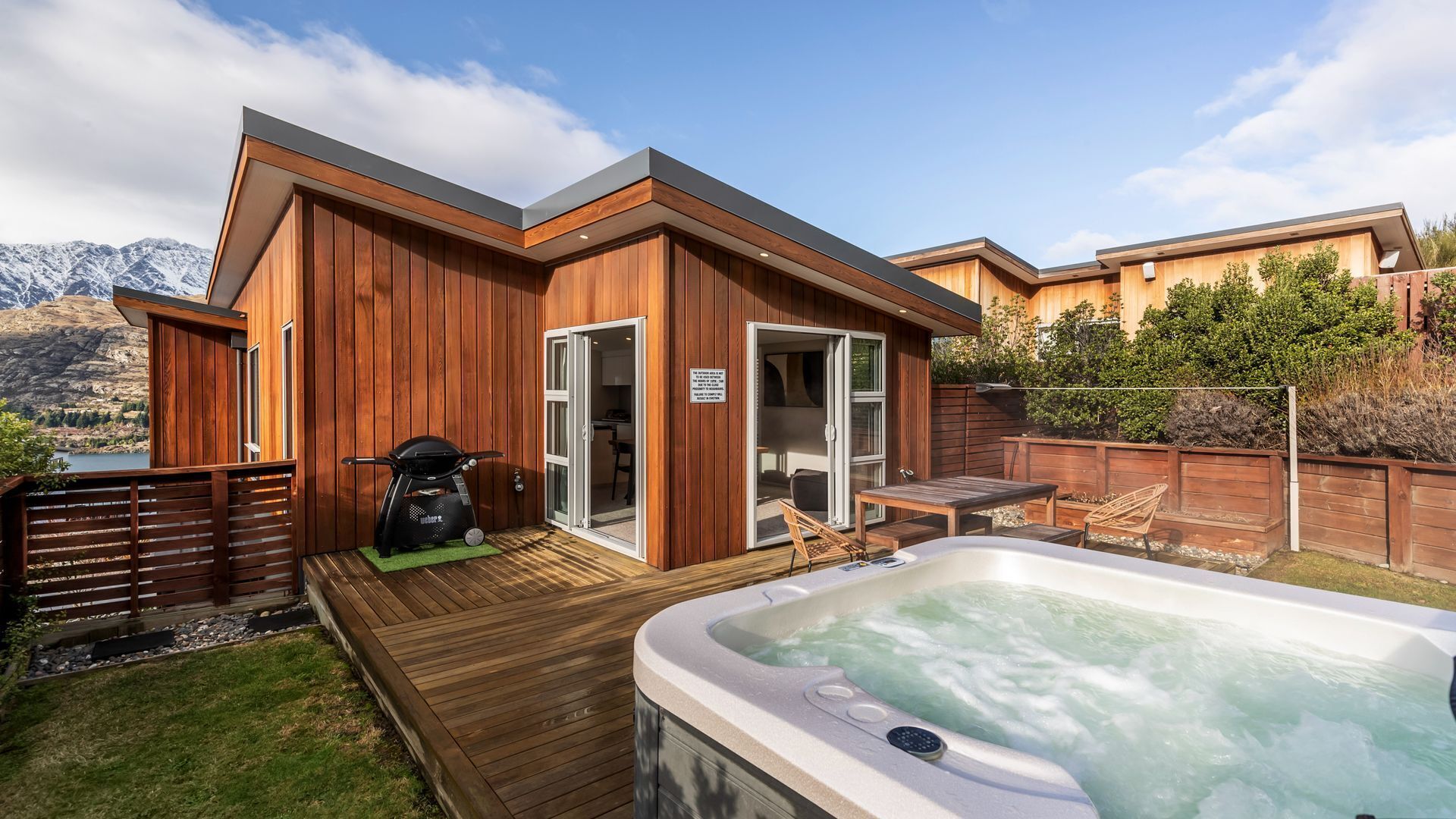 A modern outdoor hot tub area with a wooden deck, a barbecue grill, and a view of mountains.