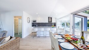 Et moderne køkken med hvide skabe, en mikrobølgeovn og en skål med frugt. Der er et spiseområde med et bord dækket til to personer, en fletstol og udsigt til det fri gennem store vinduer.