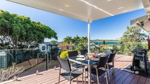 A covered outdoor dining area with a table set for two, overlooking a scenic coastal view. - Blue Waters  7 Cook St - Dog Friendly and Water Views (Salamander Bay)