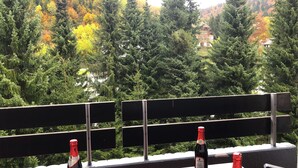 En balkong med två flaskor vin och en skål med frukt, med utsikt över en skogbeklädd sluttning där höstlöven lyser i vackra färger.