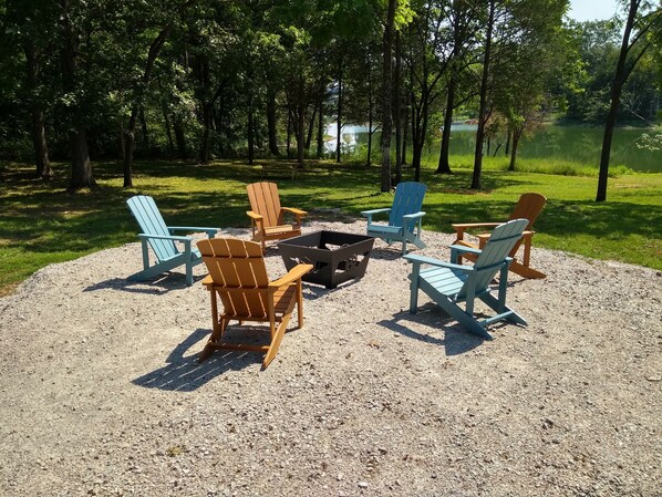 Een gebied met een vuurkuil van grind, voorzien van Adirondack-stoelen en een tafel, omgeven door bomen en een meer.