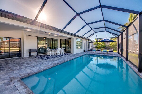 A modern indoor swimming pool with a glass enclosure, a seating area with chairs and a table, and a view of the outside.