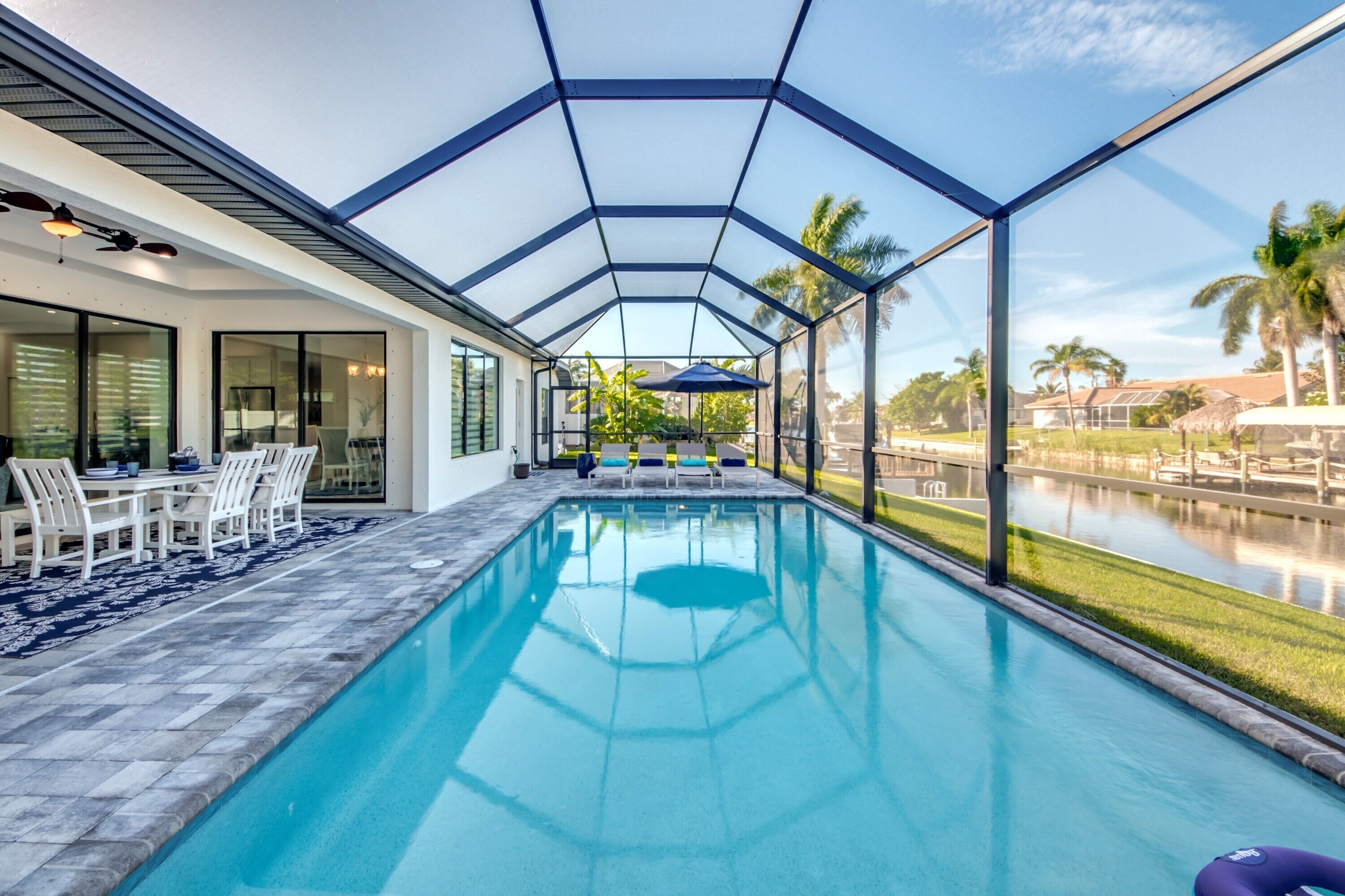 A swimming pool with a glass enclosure, a dining area with chairs and a table, and a view of a lake with palm trees.