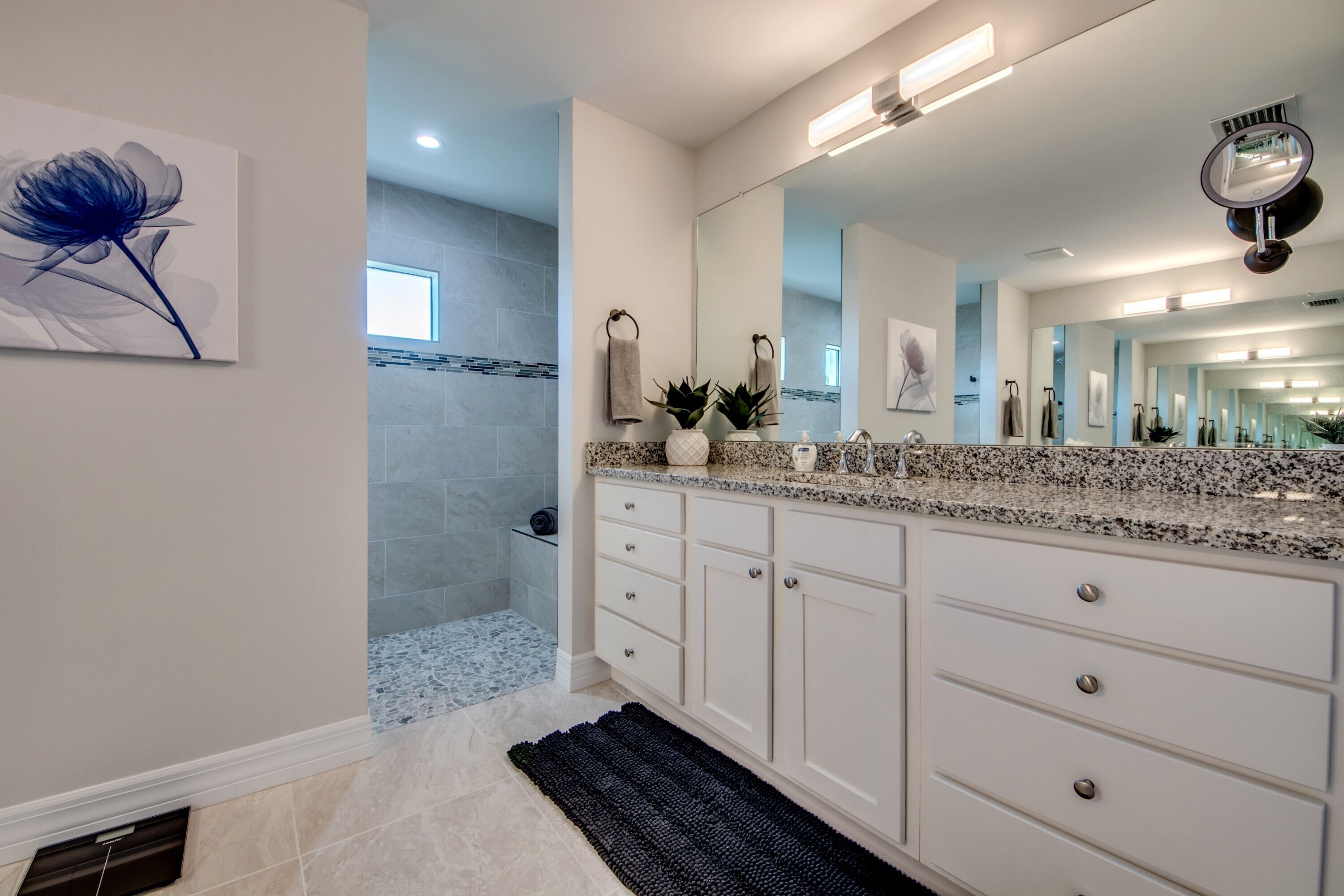 A modern bathroom with a granite countertop, white cabinetry, a large mirror, and a blue flower painting.