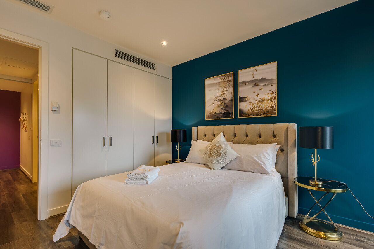A bedroom with a bed, a wardrobe, two wall-mounted artworks, a bedside lamp, and a floor lamp.