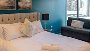 En pænt redt seng med hvide sengetæpper og dekorative puder, en natlampe, en sofa med hynder og et billede på væggen.