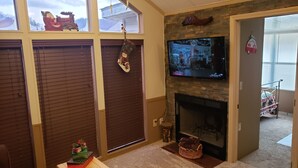 A cozy living room with a fireplace, a television mounted above it, and a window with a view of the outdoors. - Year Round Access in town Bungalow, Hot Tub, Fire Pit and Extra Parking      (Maggie Valley)