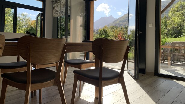 Uma área de refeições com cadeiras de madeira e uma mesa, janelas amplas com vista para as montanhas e uma porta de vidro deslizante para uma área externa.