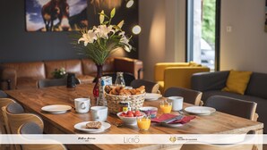 Uma área de refeições com uma mesa de madeira posta para o café da manhã, incluindo croissants, frutas e suco. Há um vaso com flores brancas, uma cesta de pão e uma pintura de burros na parede.