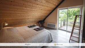Quarto com teto inclinado, cama com colcha estampada, luminária de chão e vista para uma varanda com escada de madeira.