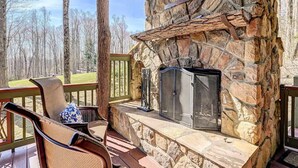 A stone fireplace with a metal screen, a wicker chair, and a bench on a wooden deck with a view of trees.