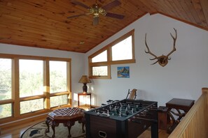 A room with a wooden ceiling, large windows, a foosball table, a tufted ottoman, and a mounted antler. - Whispering Pines Lakehouse - private dock and free zipline and railroad passes (Bryson City)