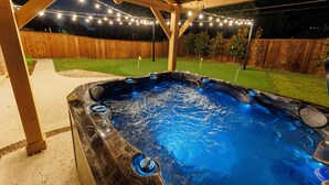 A hot tub under a wooden pergola with string lights, surrounded by a fenced yard with a grassy area and a flag. - Walk to Stadium-4 Full Bath, Hot Tub • Mini Golf • Game Room  (Arlington)