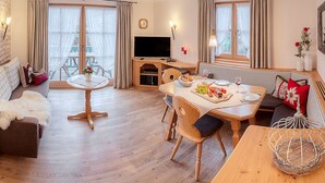 Ein gemütliches Wohnzimmer mit Essbereich, Holzmöbeln, einem Flachbildfernseher und großen Fenstern mit Vorhängen.