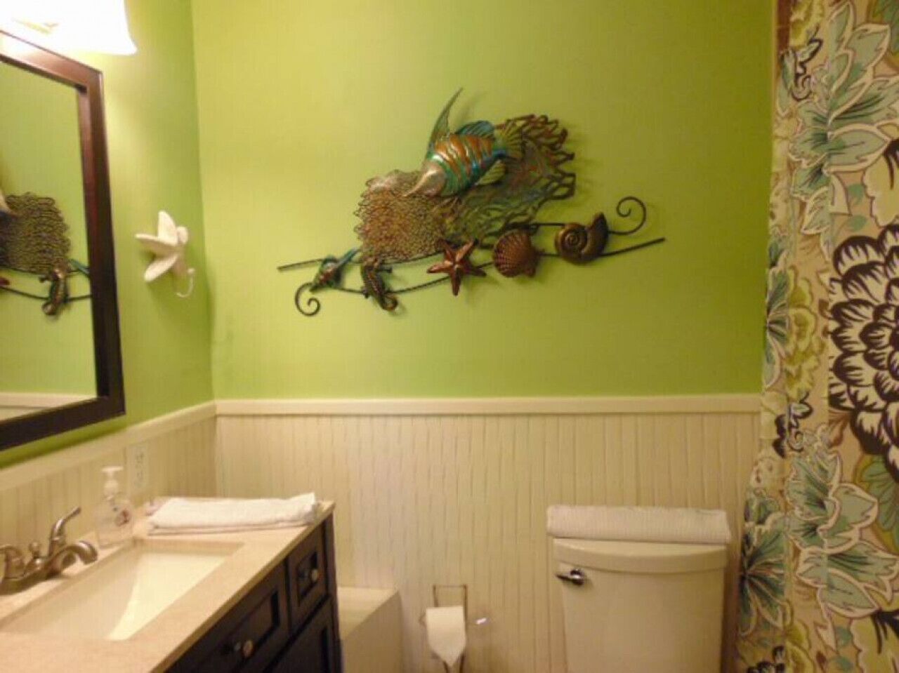 A bathroom with a green wall, a decorative fish wall art, a white toilet, and a sink with a mirror.