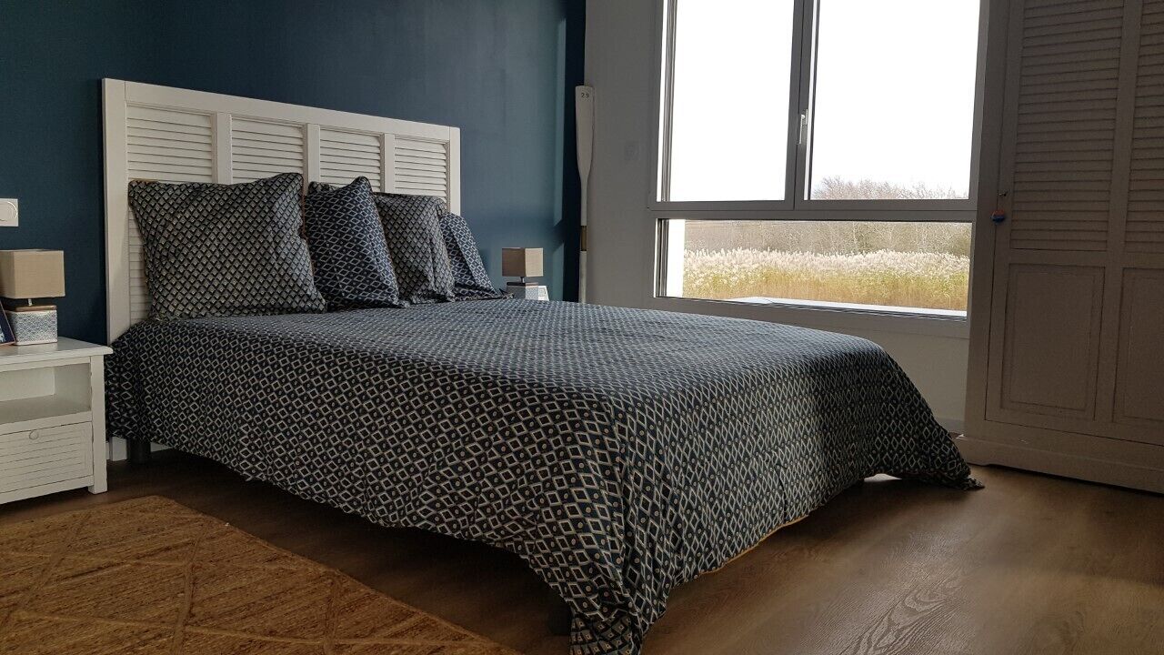 A bedroom with a patterned bedspread, a white headboard, a nightstand with a lamp, and a window with a view of a field.