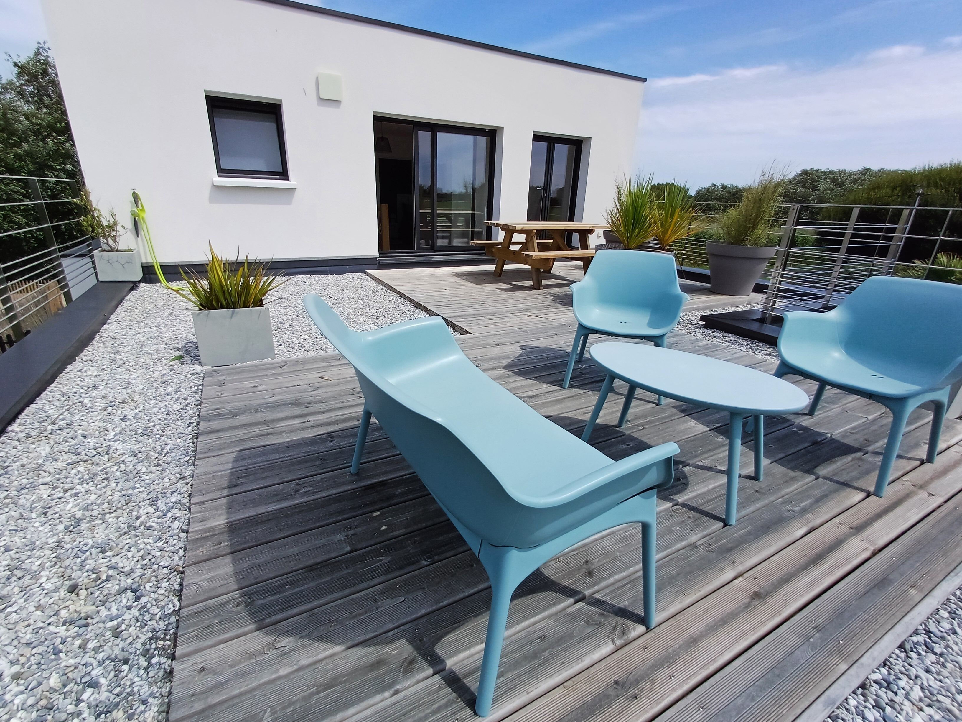 Une terrasse extérieure moderne, avec des chaises bleu clair et une table, entourée de plantes en pot et d’une table de pique-nique en bois.