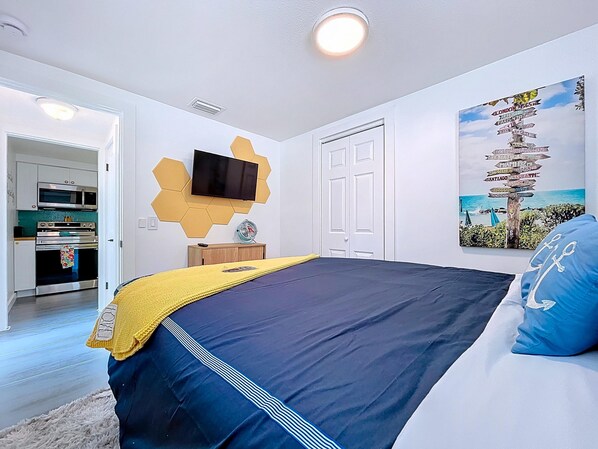 A bedroom with a bed, a TV mounted on the wall, a painting, and a kitchen visible through an open door. - Charming, Family Friendly Newly Renovated Duplex on Holmes Beach (Holmes Beach)