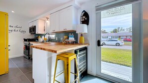 Une cuisine moderne avec des meubles blancs, un plan de travail en bois, un tabouret jaune et une vue sur la rue à travers une porte vitrée.