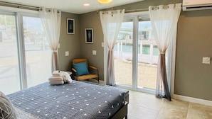 Quarto com cama, ventoinha no teto, cadeira com almofada e vista do exterior por grandes janelas.