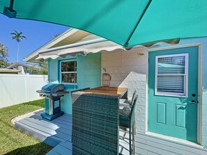 Guests can enjoy a patio with a table and grill, surrounded by palm trees and a white fence.