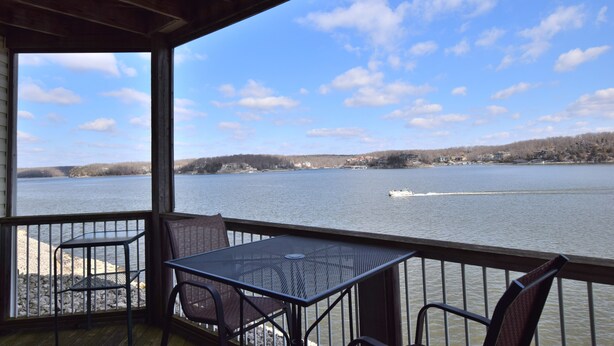 A covered outdoor area with a table and chairs facing a lake, with a view of the shoreline and a boat on the water. - Large, Beautifully Furnished -Sleeps 12,  6 beds -2 kings & 4 queens -BLDG 10 (Osage Beach)