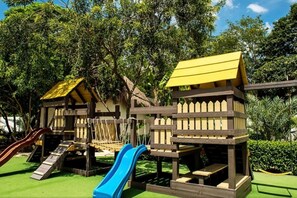 Children can play and explore in this outdoor play area featuring a playhouse and two slides surrounded by lush greenery.