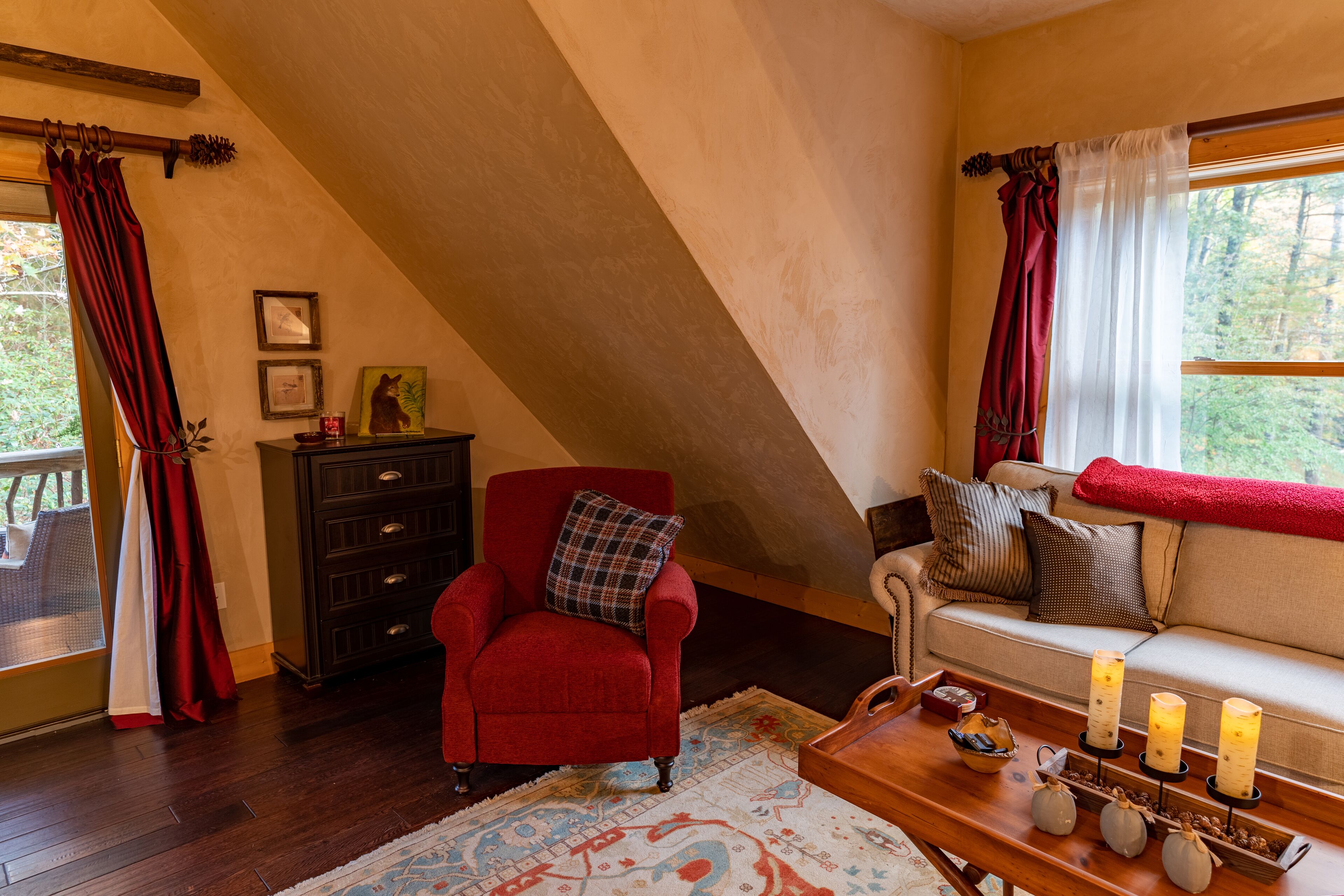 A cozy room with a red armchair, a sofa, a wooden coffee table with candles, and a view of trees through a window.