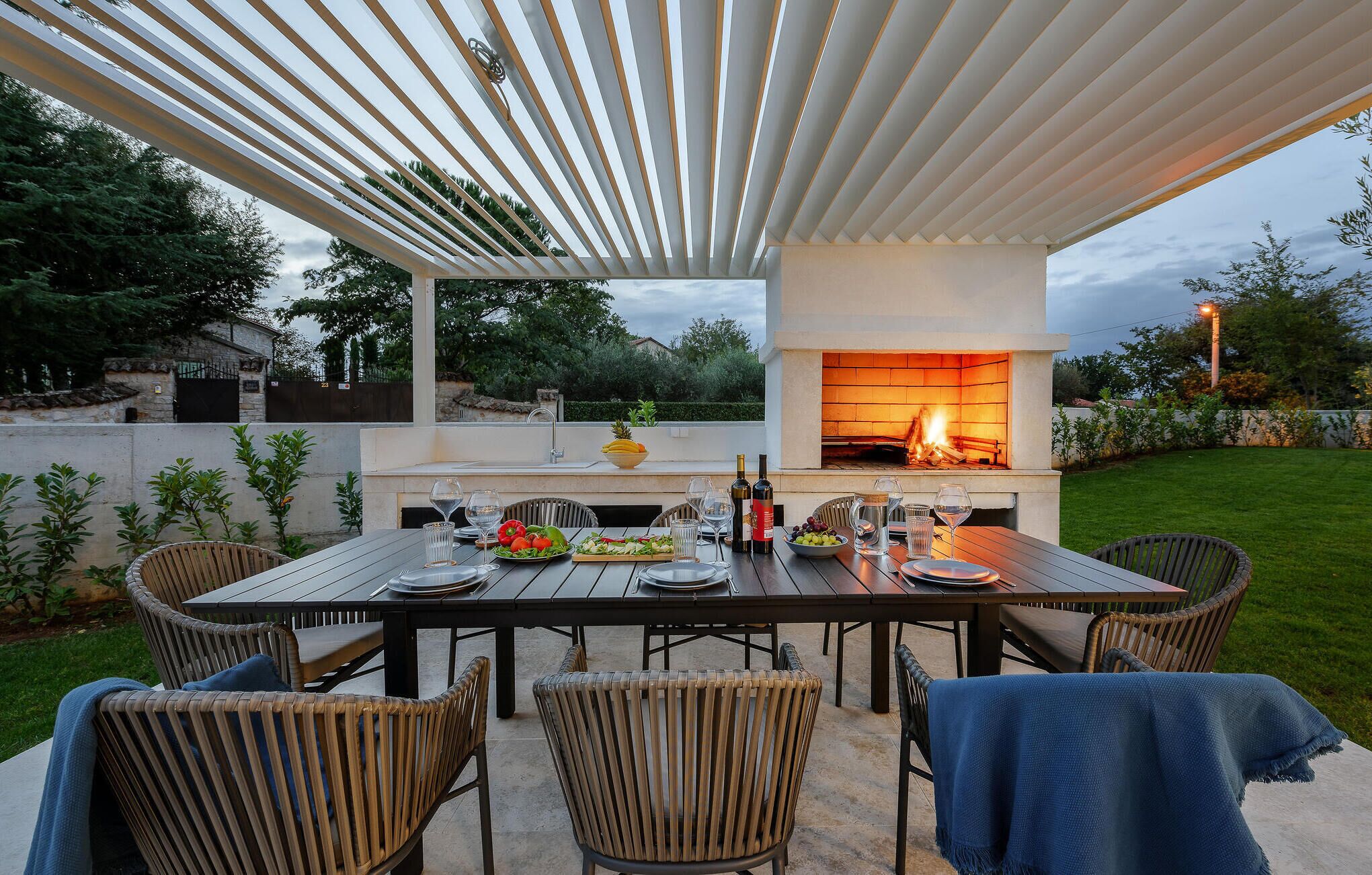 An outdoor patio with a brick fireplace, dining table, chairs, and a pergola overhead.